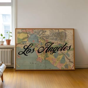 May include: A framed vintage map of Los Angeles, California, with the city name in elegant black script. The map features a muted color palette and is displayed in a room with hardwood floors and a window.