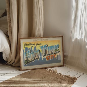 May include: A framed vintage postcard reading "Greetings from New York City." The postcard displays a skyline illustration in blue, green, and orange. The frame is a light brown colour.