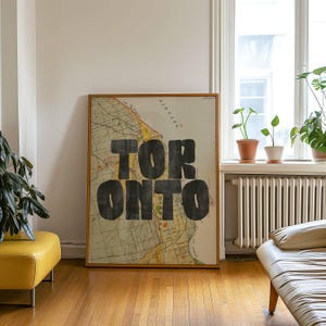 May include: A framed vintage map print of Toronto, Ontario, with the city name boldly displayed in large, black letters. The map features a detailed, aged appearance, set in a room with hardwood floors and natural light.