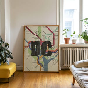 Puede incluir: Un estampado enmarcado de un mapa del metro de Washington, D.C. El mapa presenta líneas coloridas que representan las rutas del metro, con las letras negras en negrita "DC" superpuestas. El marco es de madera natural.
