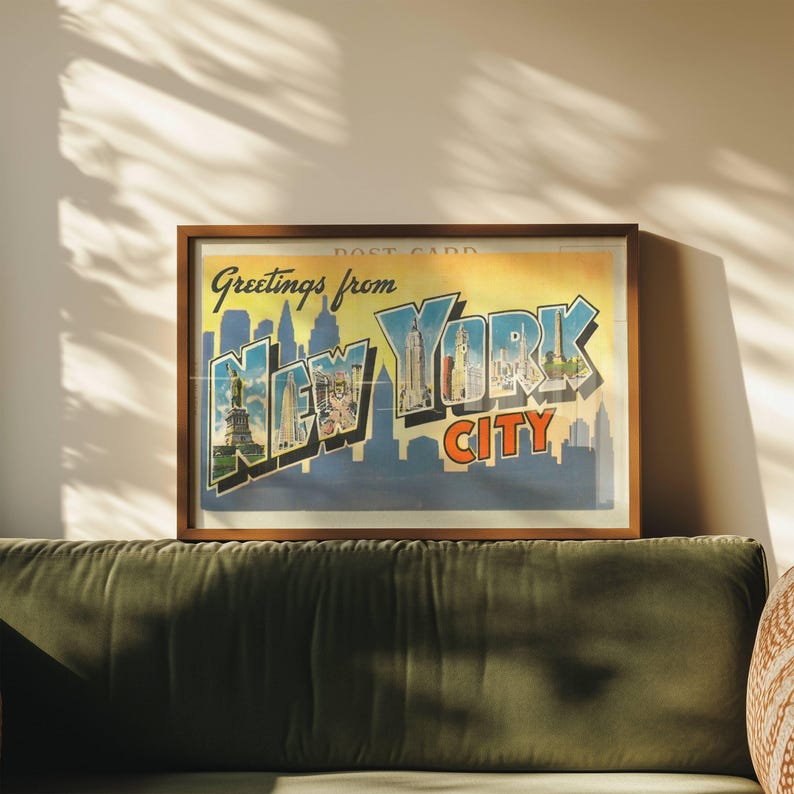 May include: A framed vintage postcard print reading "Greetings from New York City." The print displays a New York City skyline silhouette, the Statue of Liberty, and colourful text in blue and orange. The frame is made of warm brown wood.