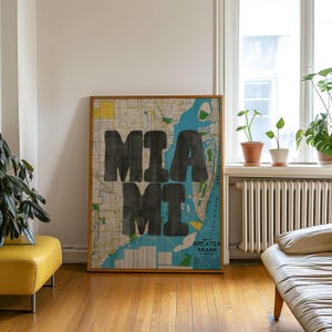 May include: A framed map of Greater Miami, Florida, with the word "MIA MI" in large, black, block letters. The map features blue, yellow, and green colors, and is displayed in a room with hardwood floors and a window.