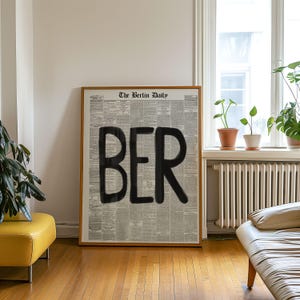 May include: A framed print featuring a newspaper page with the word "BER" in large, black letters. The newspaper is titled "The Berlin Daily". The frame is a light brown colour.