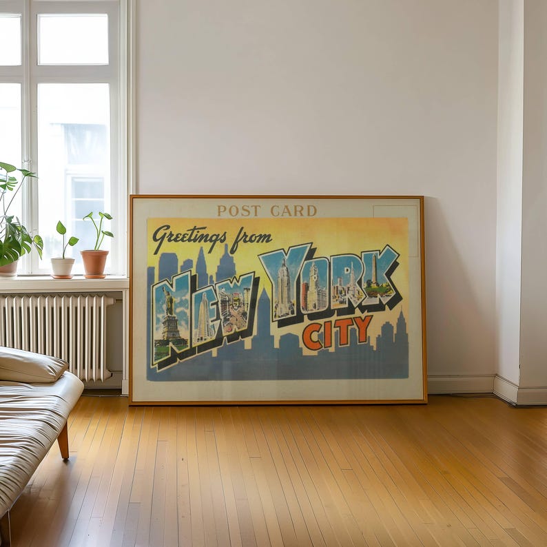 May include: A framed vintage postcard print with the words "Greetings from New York City." The artwork features a skyline silhouette and the Statue of Liberty. The print is in a wooden frame and hangs on a white wall.