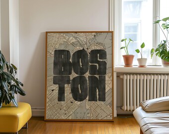 May include: A framed artwork featuring a map of Boston with the city's name boldly displayed in large, dark letters. The artwork is set against a light-colored wall, with a yellow ottoman and a window with plants in the background.