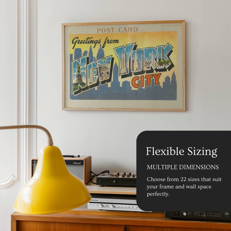 May include: A framed vintage postcard print of New York City, with the text "Greetings from New York City" in blue and yellow. The print is displayed on a white wall. A yellow lamp and a black sign that says "Flexible Sizing" are also visible.