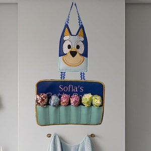 May include: A bathroom with a blue and white theme. A Bluey-themed organiser with the name "Sofia's" is mounted on the wall, holding colourful bath bombs. A light blue towel hangs below. A white toilet and sink are also visible.