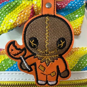 May include: Orange and brown Halloween-themed keychain featuring a cartoon character holding a lollipop. The keychain has a stitched design with a leather strap and a silver-colored clasp. The background includes a rainbow-colored sequined bow.