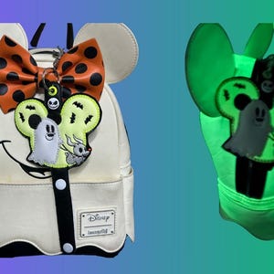 May include: Off-white backpack with a black and white smiling face design, Mickey Mouse ears, and a black and orange polka dot bow. A charm features a ghost and bat design. The backpack glows green in the dark.