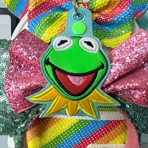 May include: A Kermit the Frog keychain with a light blue tag and a silver clasp. The keychain is attached to a white backpack with rainbow sequin bows and a rainbow striped design.