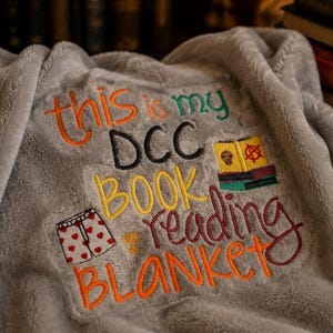 Könnte beinhalten: Eine graue Fleece-Decke, bestickt mit den Worten "this is my DCC BOOK reading BLANKET" in verschiedenen Farben. Das Design umfasst Bilder eines Buches, Unterwäsche und eines Schädels.