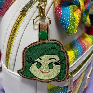 Puede incluir: Un llavero de corcho con un personaje bordado verde y amarillo, con pestañas y una sonrisa. El llavero está unido a una mochila blanca con un lazo de lentejuelas arcoíris.