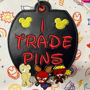 May include: A black circular faux leather tag with red embroidered text that reads I TRADE PINS, featuring two yellow embroidered mouse head silhouettes above the text and three colorful character pins attached to the bottom edge.