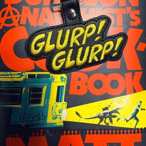 May include: Black leather keychain with the text "GLURP! GLURP!" in yellow stitching. The keychain hangs over a book cover with a train illustration and the words "THE DUNGEON ANALYST'S COOK BOOK".