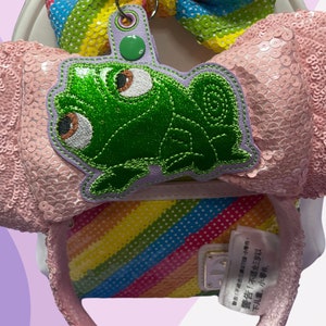 May include: Pink sequined Minnie Mouse ears headband with a rainbow striped bow and a green frog character charm. The charm has a purple border and the text "商品1 不良品は交換致しません。" is visible on the headband.