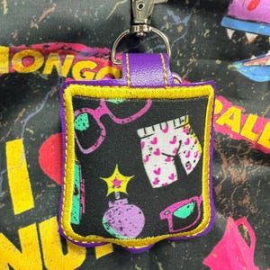 May include: A square-shaped keychain holder featuring a black fabric with a colorful pattern of pink heart-printed shorts, purple sunglasses, and a yellow star-topped bomb. The holder is trimmed with yellow stitching and a purple border.
