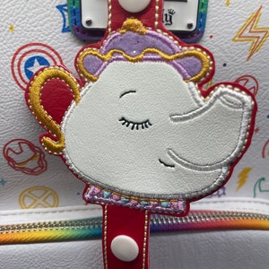 May include: A white teapot with a closed eye, embroidered on a red strap with white buttons. The strap is attached to a white background with colorful patterns.