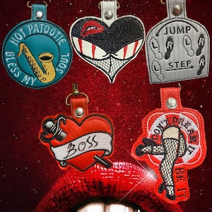 May include: Five embroidered keychains featuring various designs: a teal circle with a saxophone and text, a black heart with red lips and eyes, a grey square with dance steps, a red heart with a banner reading BOSS, and a red shape with fishnet legs.