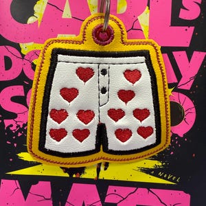 Może przedstawiać: Żółto-czarny brelok w kształcie białej bielizny z czerwonymi sercami. Brelok wisi przed książką z tytułem "Carl's Day" w różowych literach.