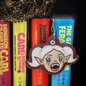 May include: A keychain featuring an embroidered vampire face with white hair, fangs, and dark eye makeup. The keychain hangs in front of a row of colorful books, including "Dungeon Crawler Carl" and "The Galaxy Fera."
