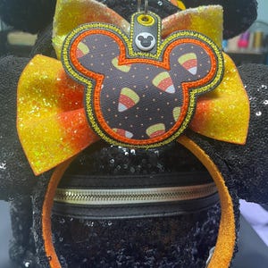 May include: Black sequined Minnie Mouse ears headband with an orange and yellow bow and a candy corn Mickey Mouse charm. The charm has a black and white candy corn pattern.