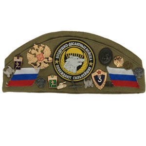 Peut inclure: Une casquette militaire verte ornée de divers épingles et patchs, dont un drapeau russe, un emblème de loup et le texte "Воздушно-десантные войска Побеждают сильнейшие".