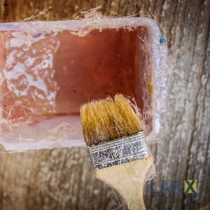 May include: A close-up of a paintbrush with a wooden handle and a silver metal band. The brush is dipped in a clear pink resin, which is in a clear plastic container. The resin is partially cured and has a textured surface.