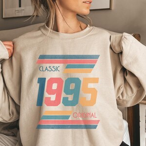 May include: A beige sweatshirt with a vintage-style graphic design featuring the year "1995" in colorful text and the words "Classic" and "Original" in black text.