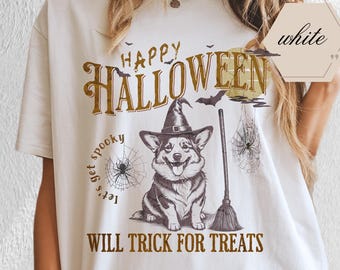 May include: White t-shirt with a Halloween-themed graphic. The design features a corgi wearing a witch hat, a broom, spiderwebs, and the text "Happy Halloween" and "Will Trick for Treats."