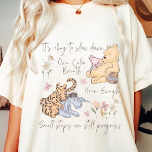 May include: Ivory t-shirt with a Winnie the Pooh design and the text "It's okay to slow down, One Calm Breath, You're Enough, Small steps are still progress." The graphic includes illustrations of Winnie the Pooh, Piglet, Tigger, and Eeyore.