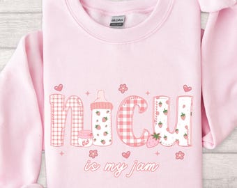 May include: Pale pink sweatshirt featuring the phrase "NICU is my jam". The text incorporates strawberry and gingham patterns, alongside a baby bottle illustration. The design includes hearts and flowers.