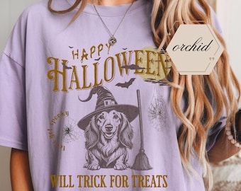 Puede incluir: Camiseta lila con un diseño de Halloween. El diseño presenta un perro salchicha con un sombrero de bruja, una escoba, telarañas y las palabras "Happy Halloween" y "Will Trick for Treats" en letras doradas.