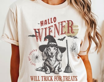 Op de afbeelding: Crèmekleurig T-shirt met een Halloween-thema. Het ontwerp toont een teckel met een heksenhoed, een bezem, spinnenwebben en de tekst "Hallo Wiener" en "Will Trick for Treats".