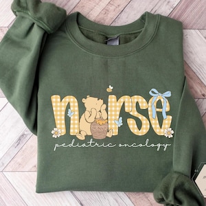 Peut inclure: Sweat-shirt vert militaire avec le mot "nurse" en imprim&eacute; vichy jaune, avec un ours de dessin anim&eacute;, un pot de miel et des accents floraux. Le texte "pediatric oncology" est en dessous.