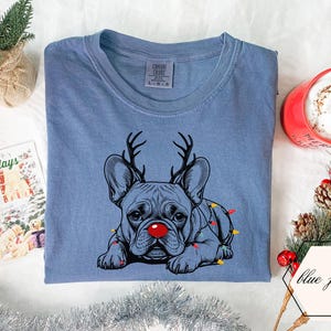 May include: Blue jean colored t-shirt featuring a French bulldog with reindeer antlers and a red nose. The dog is adorned with Christmas lights. A red mug with a white beverage and a holiday card are also in the image.