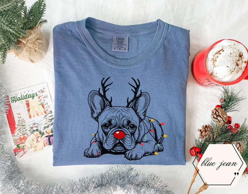 May include: Blue jean colored t-shirt featuring a French bulldog with reindeer antlers and a red nose. The dog is adorned with Christmas lights. A red mug with a white beverage and a holiday card are also in the image.