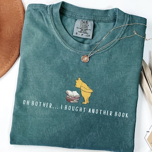 May include: Teal Comfort Colors t-shirt with the phrase "OH BOTHER... I BOUGHT ANOTHER BOOK" and a graphic of Winnie the Pooh. A gold necklace and a brown hat are also visible.