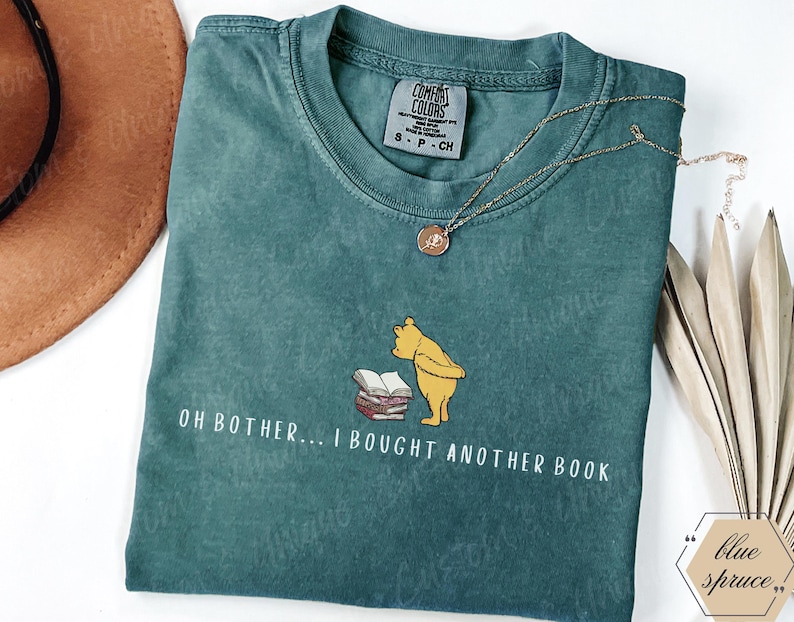 May include: Teal Comfort Colors t-shirt with the phrase "OH BOTHER... I BOUGHT ANOTHER BOOK" and a graphic of Winnie the Pooh. A gold necklace and a brown hat are also visible.