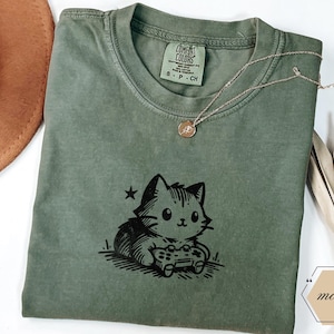 May include: A moss green Comfort Colors t-shirt featuring a black graphic of a cat holding a video game controller. A gold necklace with a pendant rests on the shirt. The shirt has a small tag on the collar.