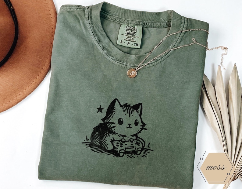 May include: A moss green Comfort Colors t-shirt featuring a black graphic of a cat holding a video game controller. A gold necklace with a pendant rests on the shirt. The shirt has a small tag on the collar.