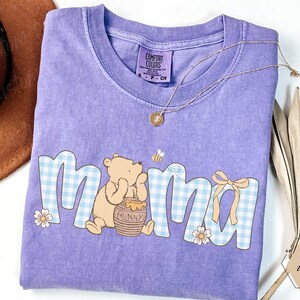 May include: A lilac Comfort Colors t-shirt featuring the word "Mama" in a blue gingham pattern with a Winnie the Pooh graphic. The design includes a honey pot, a bee, and a bow. A gold necklace and a brown hat are also visible.
