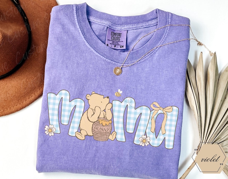 May include: A lilac Comfort Colors t-shirt featuring the word "Mama" in a blue gingham pattern with a Winnie the Pooh graphic. The design includes a honey pot, a bee, and a bow. A gold necklace and a brown hat are also visible.