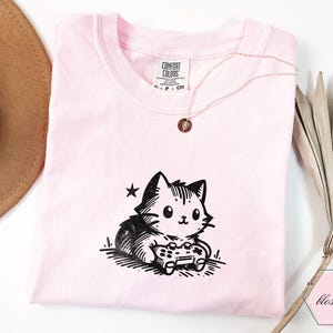 May include: A light pink t-shirt featuring a black and white cartoon cat holding a video game controller. The shirt has a crew neck and short sleeves. A gold necklace with a small pendant is on the shirt.