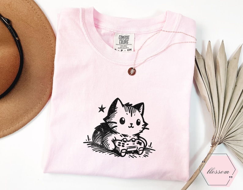 May include: A light pink t-shirt featuring a black and white cartoon cat holding a video game controller. The shirt has a crew neck and short sleeves. A gold necklace with a small pendant is on the shirt.