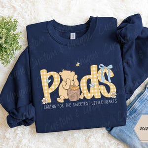May include: Navy blue sweatshirt with the word "Pods" in yellow gingham letters, featuring a cartoon bear and honey pot. The text "Caring for the sweetest little hearts" is below the word "Pods".