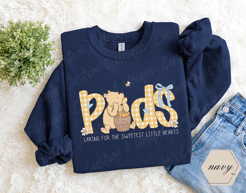 May include: Navy blue sweatshirt with the word "Pods" in yellow gingham letters, featuring a cartoon bear and honey pot. The text "Caring for the sweetest little hearts" is below the word "Pods".