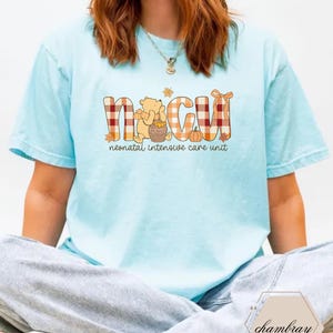 May include: Light blue t-shirt with the letters "NICU" in a plaid design, featuring a cartoon bear and pumpkin. The text "neonatal intensive care unit" is below the letters. The shirt is a casual style.