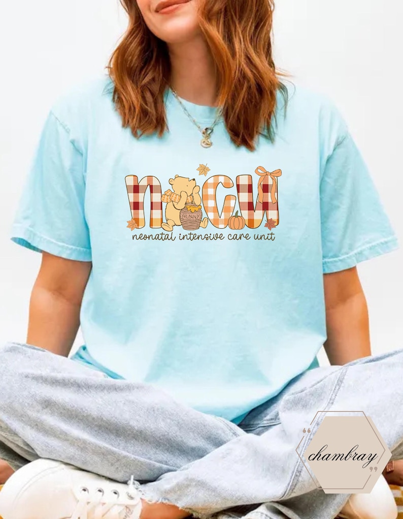 May include: Light blue t-shirt with the letters "NICU" in a plaid design, featuring a cartoon bear and pumpkin. The text "neonatal intensive care unit" is below the letters. The shirt is a casual style.