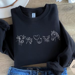 Op de afbeelding: Zwart sweatshirt met een witte omtrek van een koe, geit, stethoscoop, varken en paard. Het ontwerp is gecentreerd op de voorkant van het sweatshirt.
