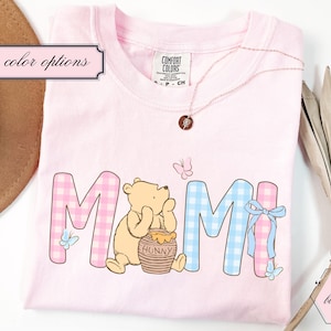 Könnte beinhalten: Ein hellrosa T-Shirt mit dem Wort "MOM" in einem Vichy-Muster, mit Winnie Puuh und einem Honigtopf. Das Shirt hat auch Schmetterlinge und eine Halskette.